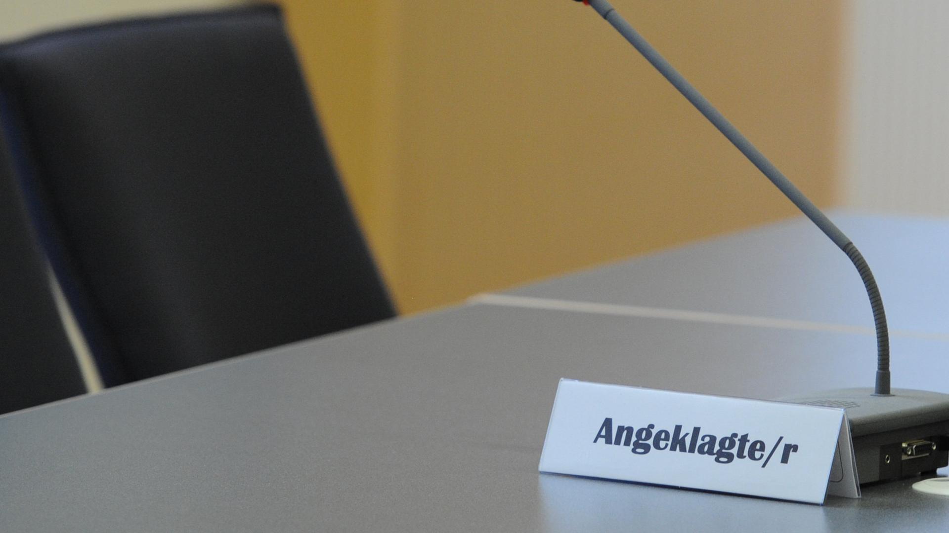 Der Sitzplatz des Angeklagten in einem Gerichtssaal. Auf dem Tisch vor dem Stuhl steht ein Mikrofon und ein Schild mit der Aufschrift "Angeklagte/r".