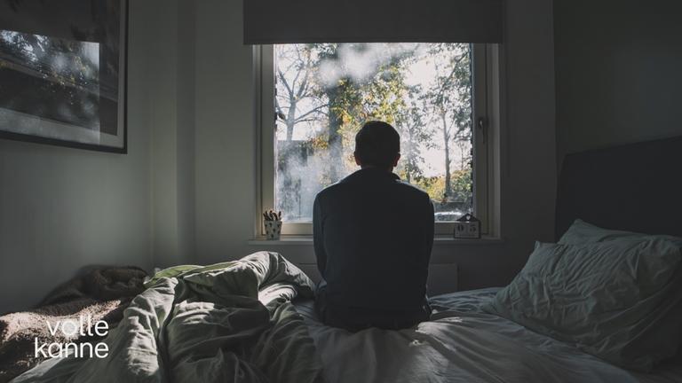 Ein Mann sitzt auf einem Bett vor einem Fenster und schaut hinaus