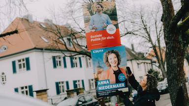Zwei Wahlplakate an einem Laternenmast, darauf sind eine Frau und ein Mann zu sehen. 