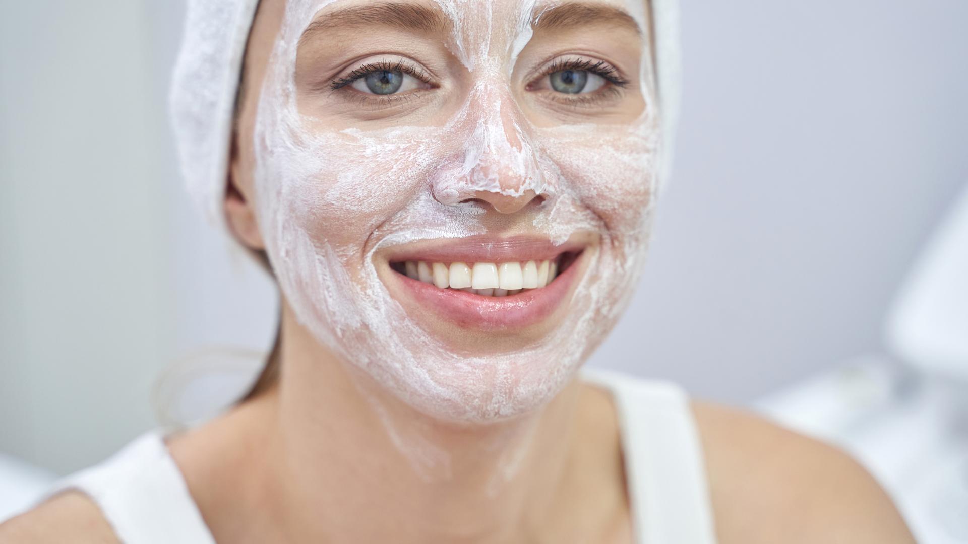 Eine Frau schaut mit einer Gesichtsmaske im Gesicht lächelnd in die Kamera. Um ihre Haare hat sie ein weißes Handtuch gewickelt.