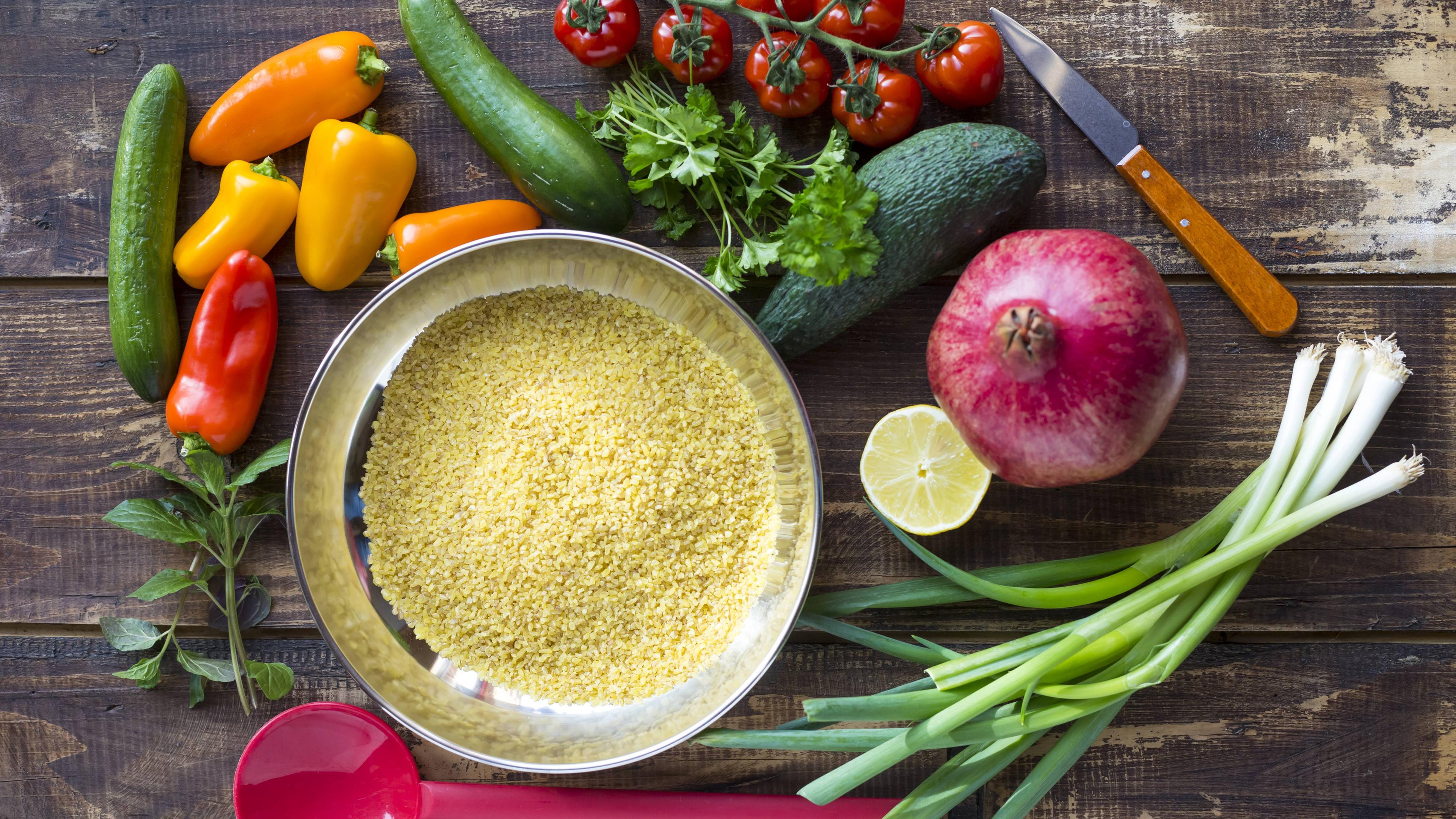 Zutaten für einen Bulgursalat auf einem Tisch: Bulgur, Paprika, Zucchini, Tomaten, Avocado, Granatapfel, Frühlingszwiebeln, Zitrone, Kräuter