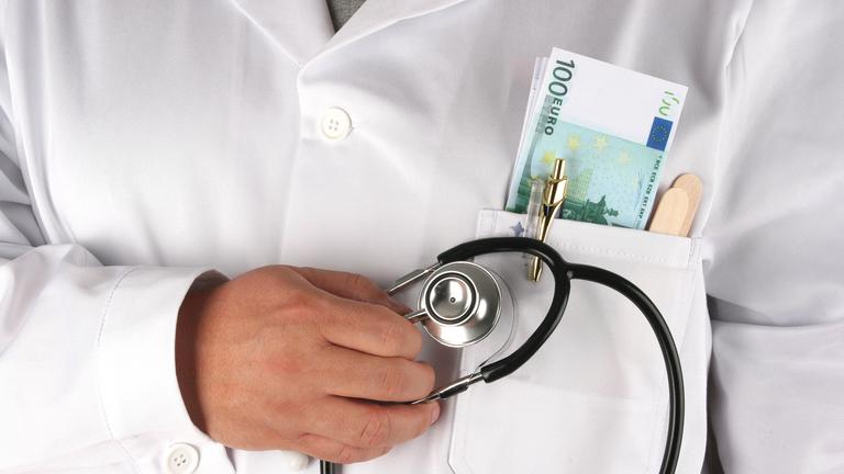In dem weißen Kittel eines Arztes befindet sich in der Brusttasche neben einem Stethoskop noch ein Bündel mit Geldscheinen.