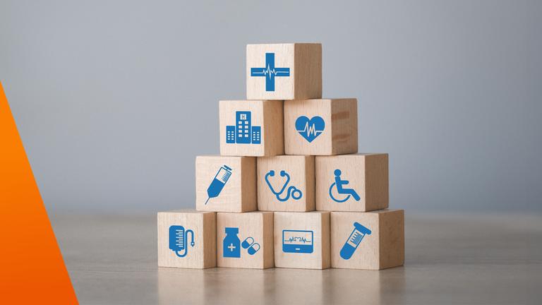 Auf einem Tisch sind Holzklötze zu einer kleinen Pyramide aufgebaut. Die Klötze tragen blaue medizinische Symbole wie EKG, Spritze, Tabletten, Rollstuhl und Klinikgebäude. Das Bild visualisiert die vielfältigen Elemente des Gesundheitswesens und deren Zusammenspiel.