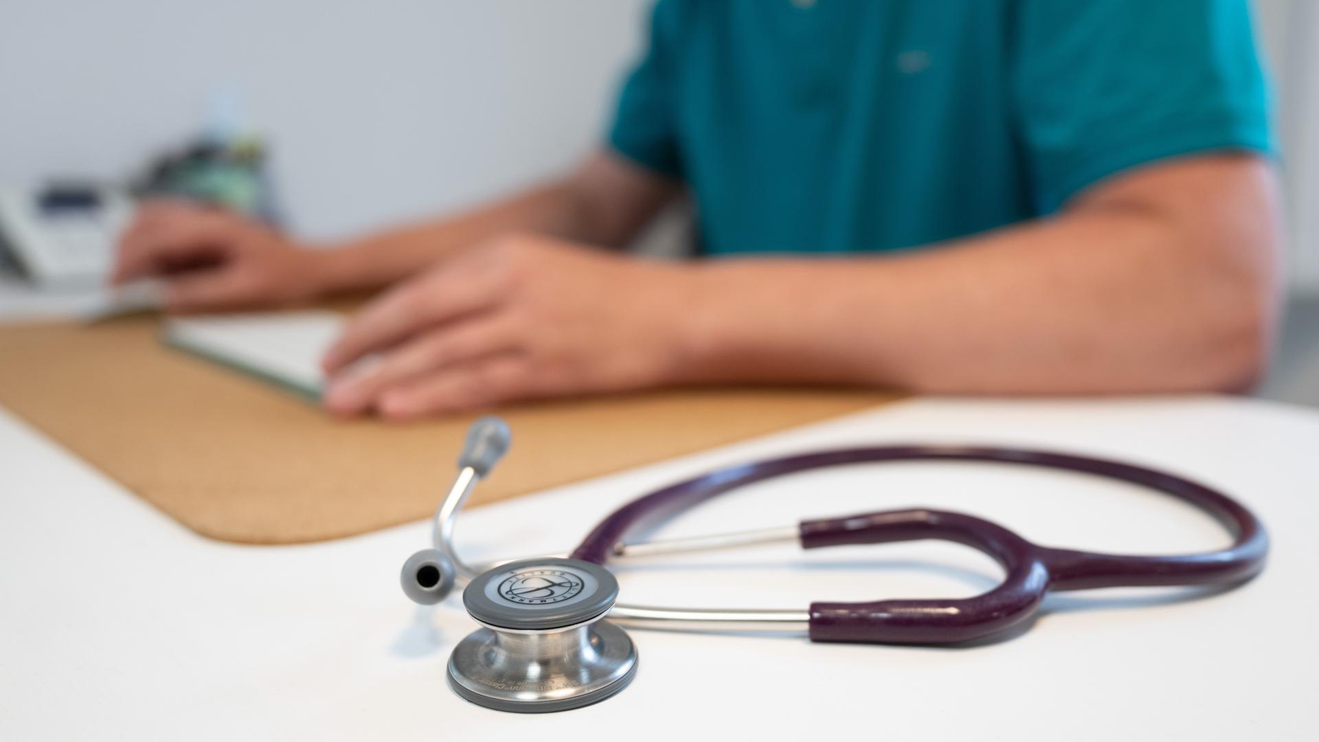 Symbolbild: Ein Hausarzt sitzt in einem Sprechzimmer seiner Hausarztpraxis an einem Schreibtisch neben einem Stethoskop und arbeitet am Computer.