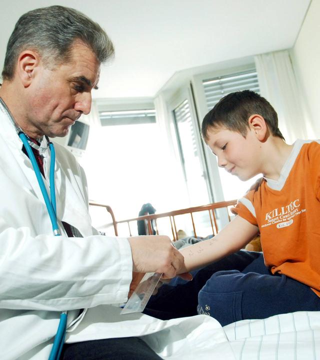 Ein Arzt in Kittel zeigt einem Jungen, der auf einem Krankenhausbett sitzt, wie ein Allergietest funktioniert.