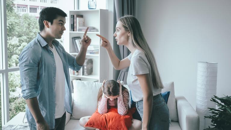 Ein Mann und eine Frau streiten sich, während ein Mädchen auf dem Sofa hinter ihnen sitzt und sich die Ohren zuhält.