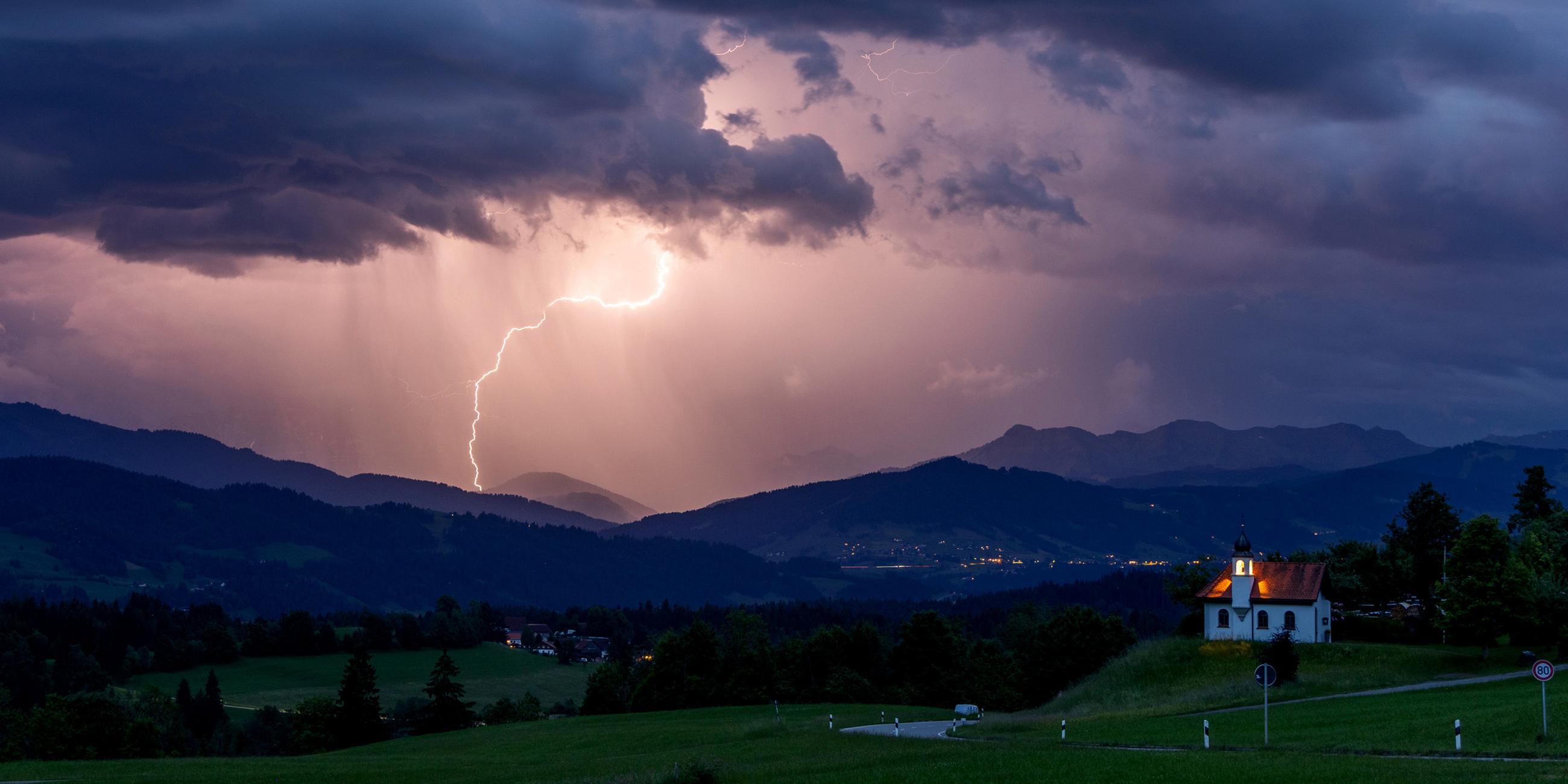 Blitze entladen sich über den Alpen während eines aufziehenden Gewitters am Abend hinter der St. Hubertus Kapelle in Scheidegg am 01.06.2025.