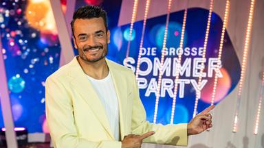 Moderator Giovanni Zarrella lächelt in die Kamera und zeigt mit beiden Zeigefingern auf das Sendungslogo "Die große Sommerparty".