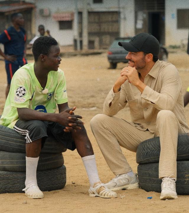 Moderator Giovanni Zarrella sitzt mit einem Jugendlichen auf gestapelten Reifen in einem Armenviertel von Abidjan 
