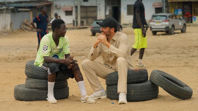 Moderator Giovanni Zarrella sitzt mit einem Jugendlichen auf gestapelten Reifen in einem Armenviertel von Abidjan 