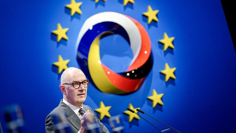 Auf dem Bild ist das Logo der Konferenz zu sehen. Davor steht ein Mann auf der Bühne und spricht ins Mikrofon.