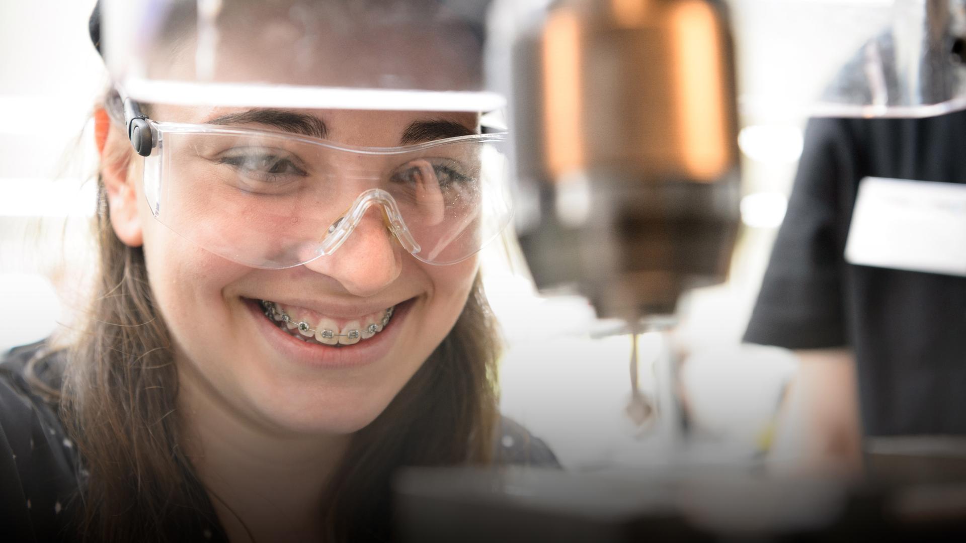 Ein junges Mädchen trägt einen Helm und eine Schutzbrille und bedient lachend eine Fräse.