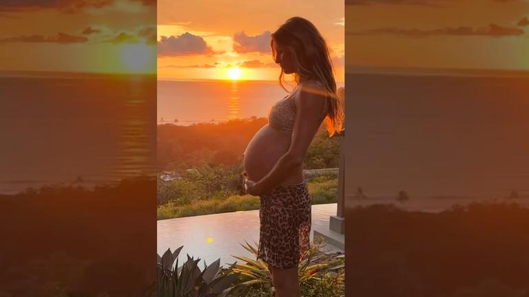 Die schwangere Gisele Bündchen steht vor einem Sonnenuntergang.