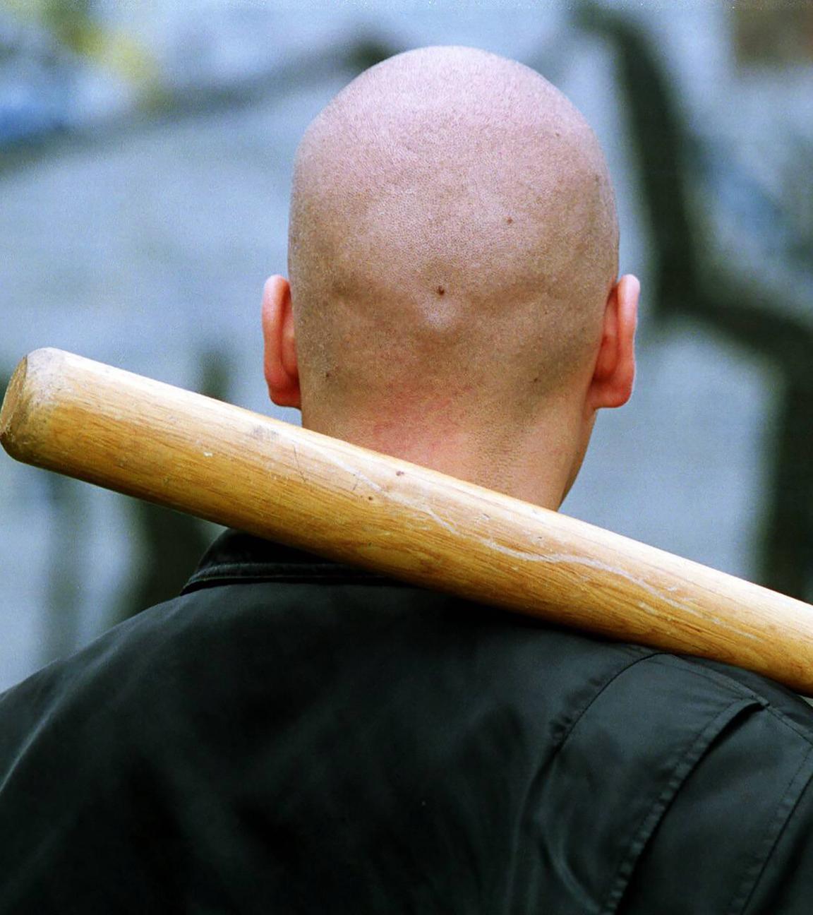 Rückansicht eines Mannes mit kahlgeschorenem Kopf, Bomberjacke und einem Baseballschläger, den er sich über die Schulter gelegt hat.