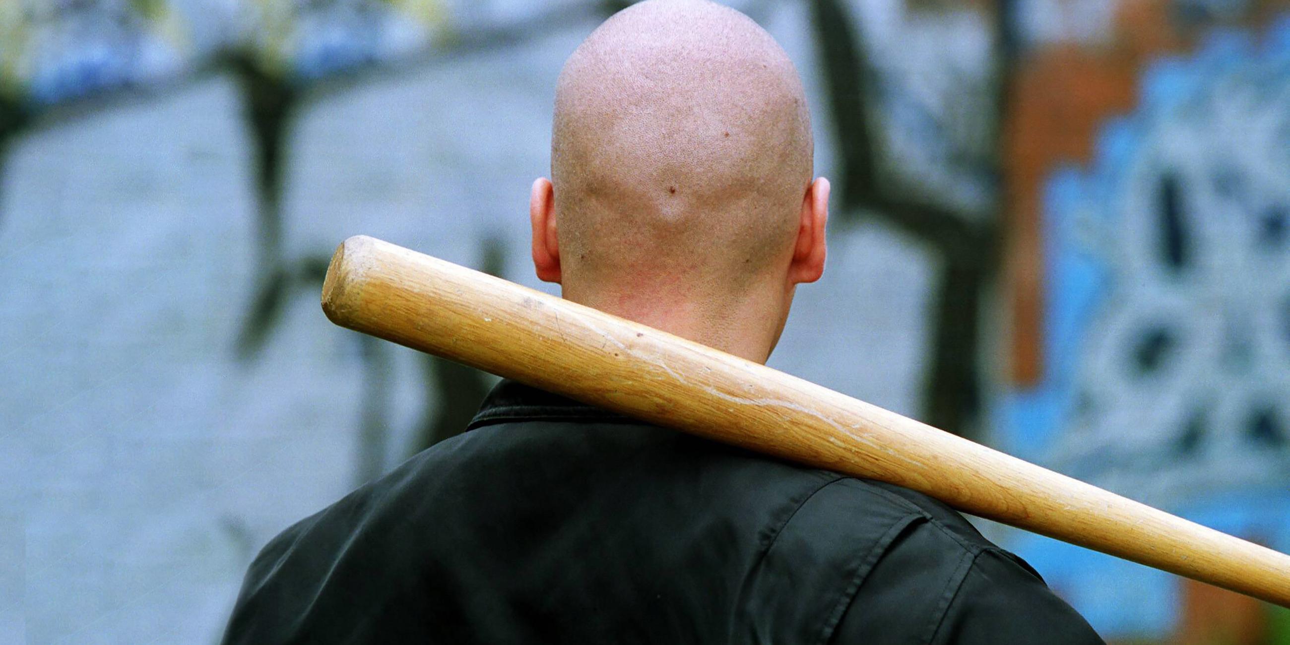 Rückansicht eines Mannes mit kahlgeschorenem Kopf, Bomberjacke und einem Baseballschläger, den er sich über die Schulter gelegt hat.