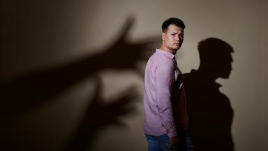 Max Leon steht in Jeans und rosa Hemd seitlich vor einer Wand. Hinter ihm ist sein Schatten an der Wand und Schattenhände, die nach ihm greifen wollen.