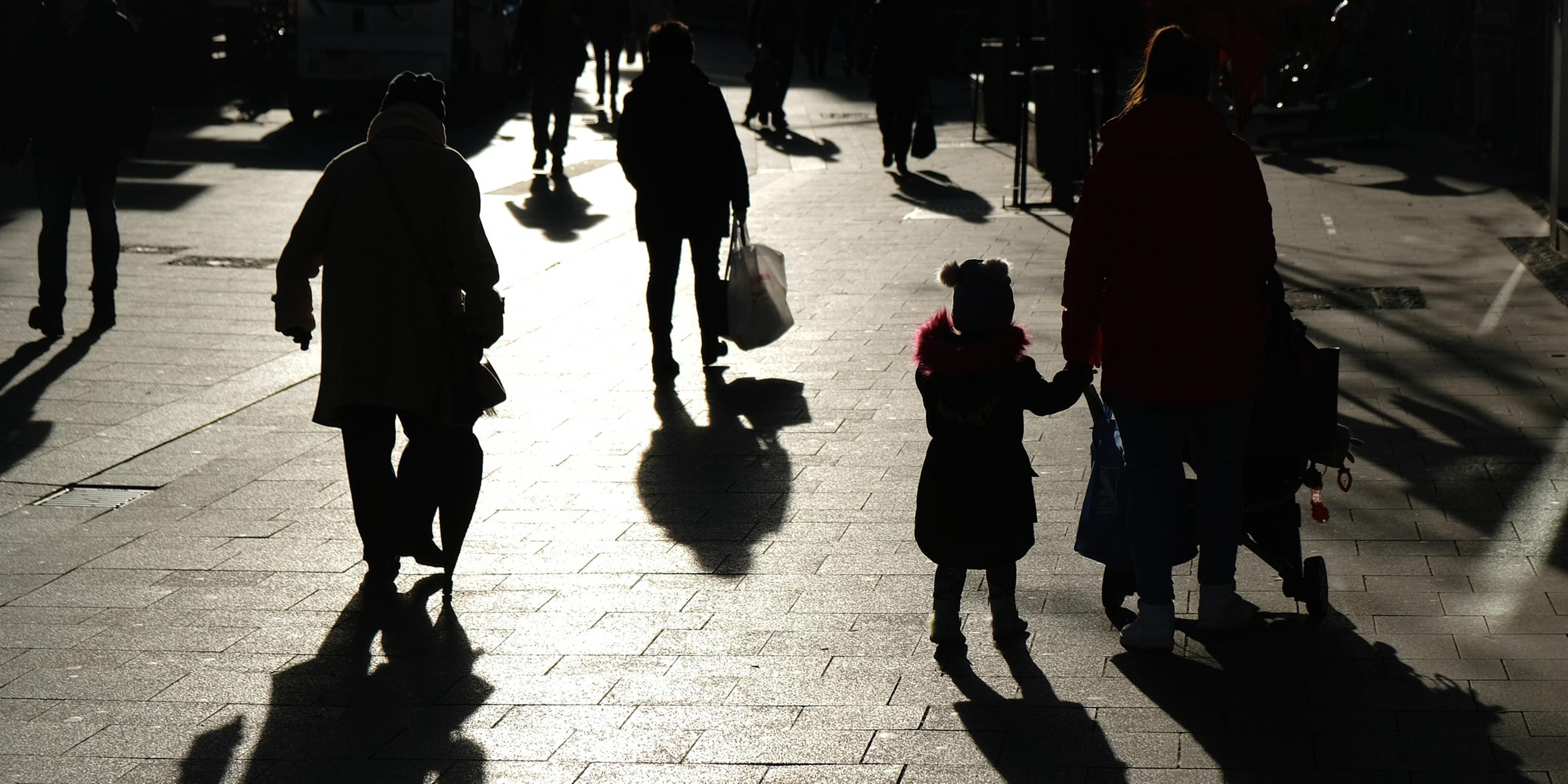 Passanten mit Tüten gehen während des Lockdowns durch eine Fußgängerzone und sind dabei als Silhouette und Schatten zu sehen.