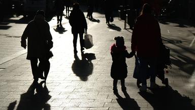 Passanten mit Tüten gehen während des Lockdowns durch eine Fußgängerzone und sind dabei als Silhouette und Schatten zu sehen.