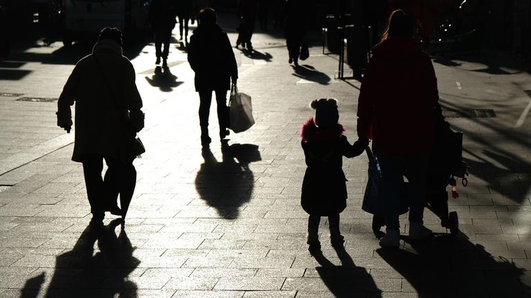 Passanten mit Tüten gehen während des Lockdowns durch eine Fußgängerzone und sind dabei als Silhouette und Schatten zu sehen.