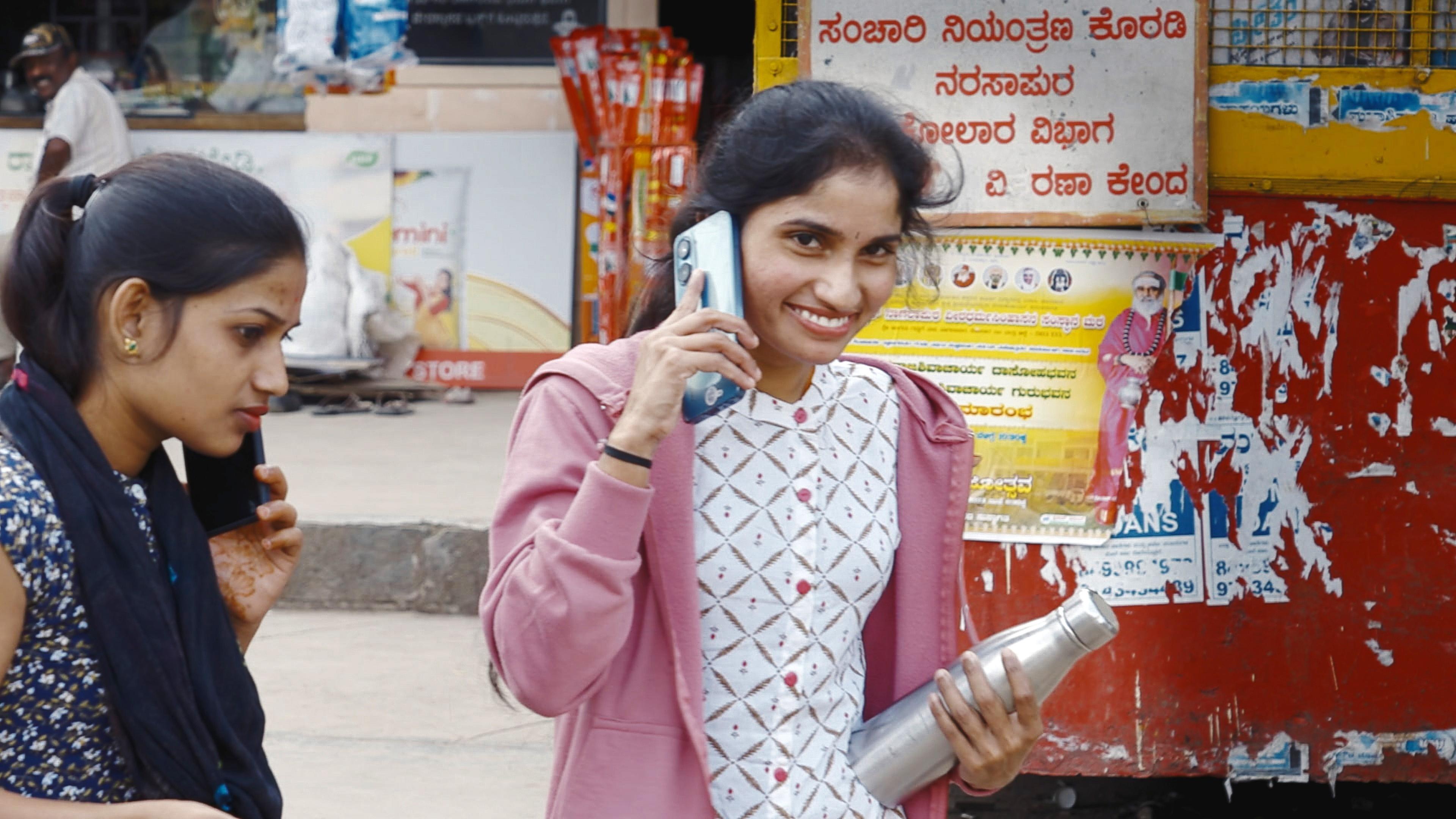 Zwei junge Frauen telefonieren mit ihren Smartphones auf der Straße in der Provinz Bangalore, eine lächelt in die Kamera.