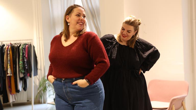 Nicole, 39 Jahre alt, steht in der Bildmitte. Sie trägt Jens und einen roten Pullover, ihre dunkelblonden Haare sind halblang und am Oberkopf nach hinten gekämmt. Sie hat runde Ohrringe, ihre Lippen sind rot geschminkt. Hinter ihr steht eine junge blonde Frau, Mitte 20. Diese trägt ein elegantes schwarzes Mini-Kleid mit Puffärmeln. Sie guckt auf die andere Frau, die offensichtlich die Kleidungsstücke probiert und sich im Spiegel betrachtet, der jedoch außerhalb des Bildes ist.