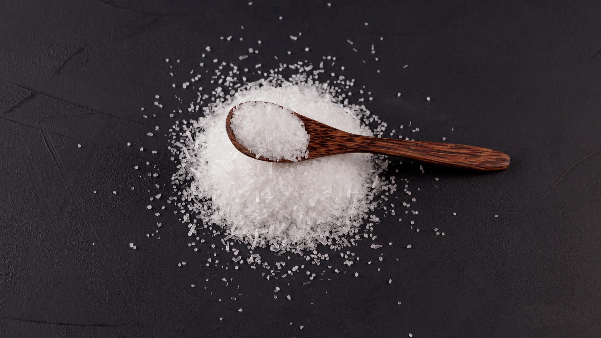 Glutaminsäuremononatriumsalz vor einem schwarzen Hintergrund mit einem Holzlöffel darauf.