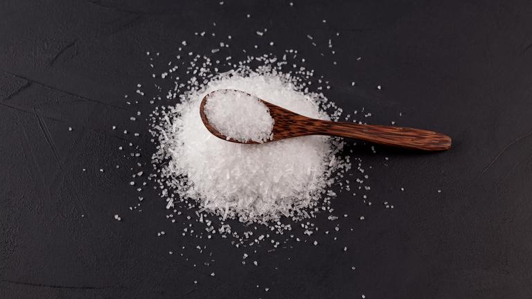 Glutaminsäuremononatriumsalz vor einem schwarzen Hintergrund mit einem Holzlöffel darauf.