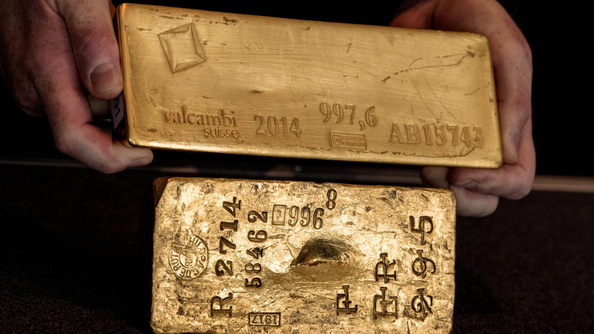 Zwei Goldbarren werden vor einem schwarzen Hintergrund in den Händen gehalten.
