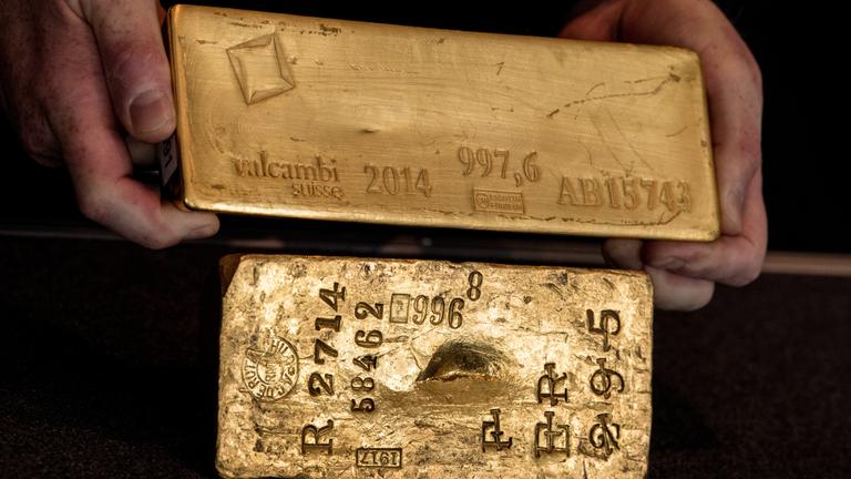 Zwei Goldbarren werden vor einem schwarzen Hintergrund in den Händen gehalten.