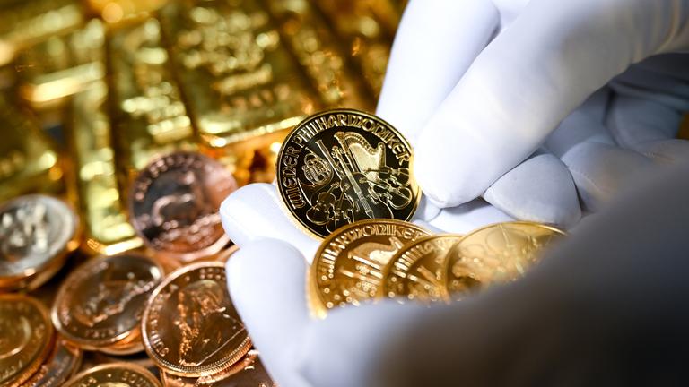 Goldmünzen werden in die Kamera gehalten, im Hintergrund weitere Münzen und Goldbarren.