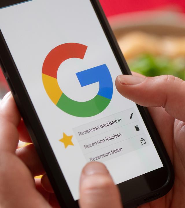 Eine Hand hält vor einem gedeckten Esstisch ein Smartphone mit dem Google-Logo und dem Menüpunkt eine Rezension zu bearbeiten, zu löschen oder zu teilen.