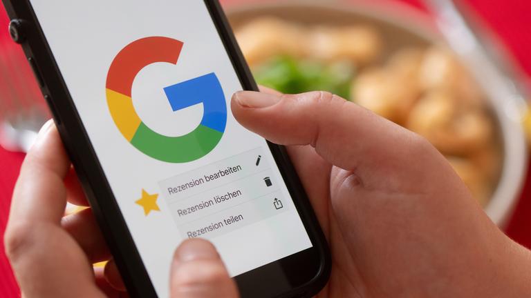 Eine Hand hält vor einem gedeckten Esstisch ein Smartphone mit dem Google-Logo und dem Menüpunkt eine Rezension zu bearbeiten, zu löschen oder zu teilen.