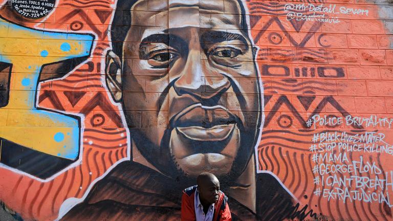 Ein Mann sitzt vor einem Graffiti von George Floyd.