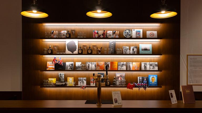 Eine Theke mit Zapfhahn und dahinter ist ein Bücherregal, in dem verschiedene Bilder, Bücher, Parfums und auch Flaggen stehen. Von oben hängen drei Lampen runter ins Bild.