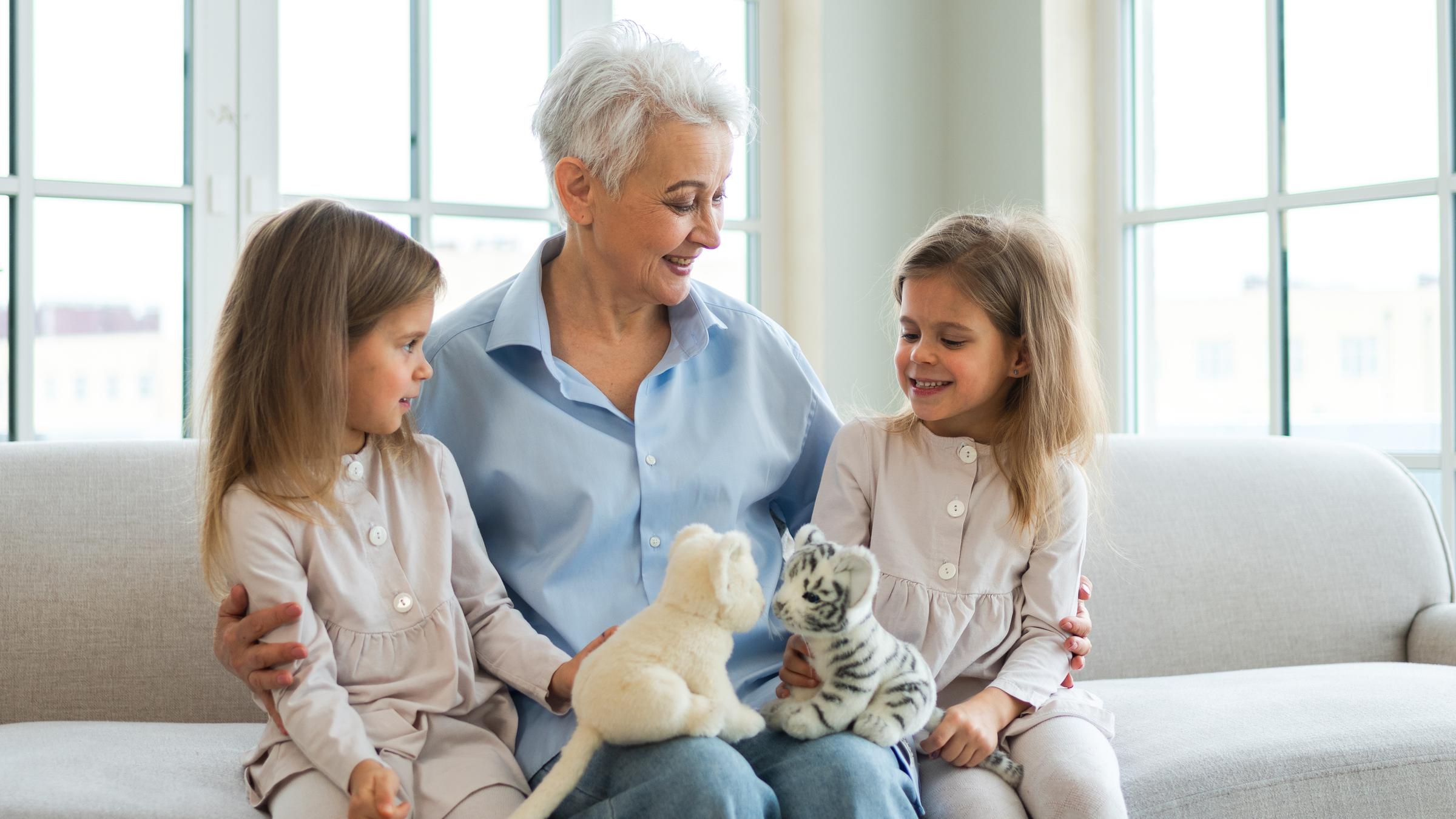 Eine ältere Frau sitzt zwischen zwei Mädchen auf einem grauen Sofa, auf ihrem Schoß liegen zwei Plüschkatzen.