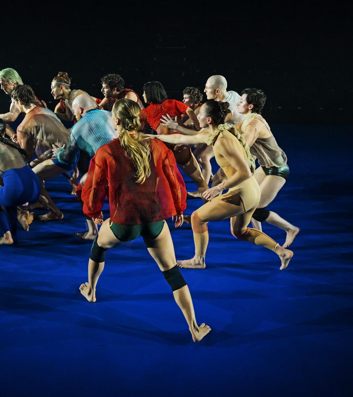 15 leicht und bunt bekleidete Tänzer auf einer blauen Bühne laufen in einer Gruppe, berühren den Vordermann, oder die Vorderfrau mit einer Hand und blicken alle in eine Richtung.