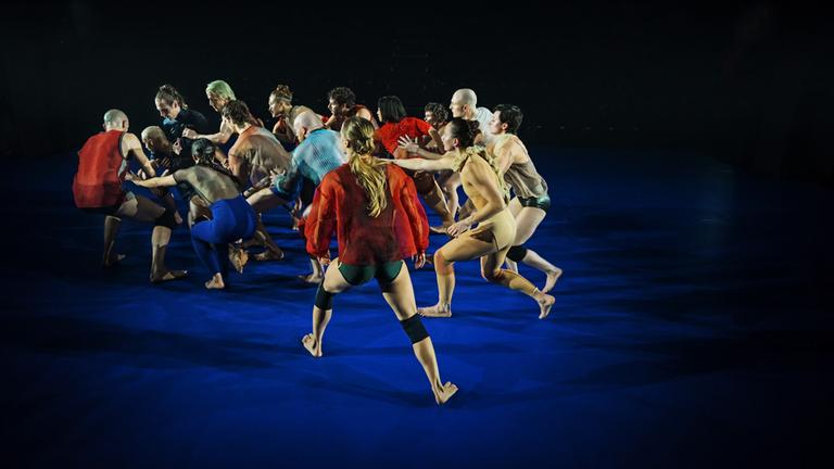 15 leicht und bunt bekleidete Tänzer auf einer blauen Bühne laufen in einer Gruppe, berühren den Vordermann, oder die Vorderfrau mit einer Hand und blicken alle in eine Richtung.