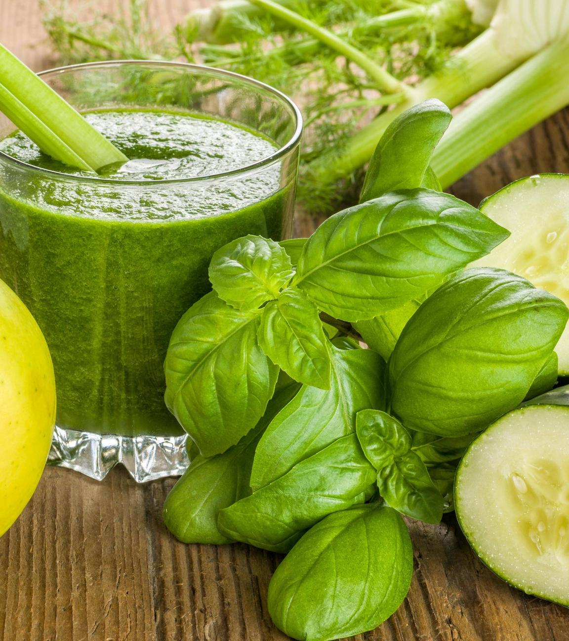 Grüner Drink im Glas, davor liegen ein Apfel, frischer Basilikum und eine angeschnittene Gurke 
