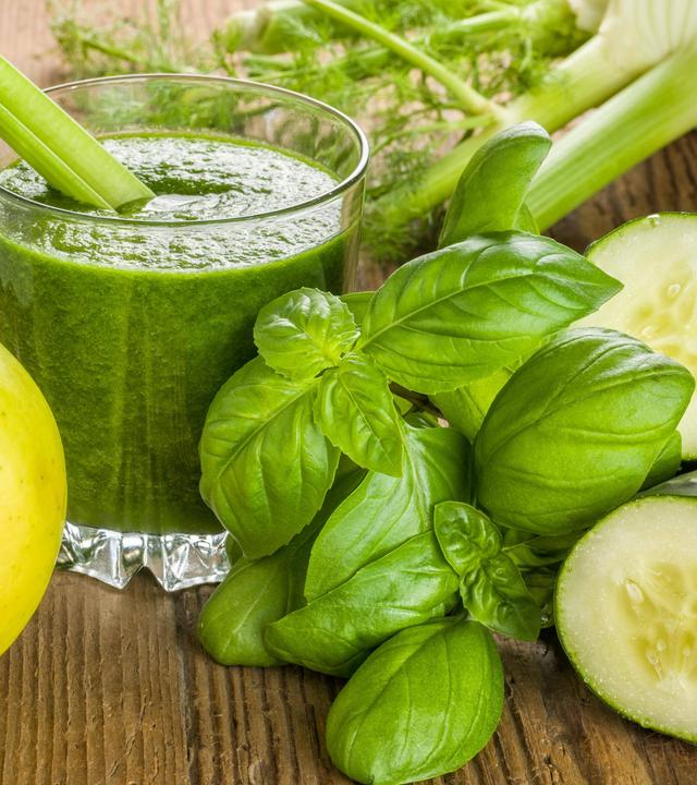 Grüner Drink im Glas, davor liegen ein Apfel, frischer Basilikum und eine angeschnittene Gurke 
