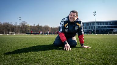 Greenkeeper Sebastian Breuing kniet in schwarzer Jacke mit DFB-Logo auf einem Fußballfeld und stützt seine Hände auf den Rasen.