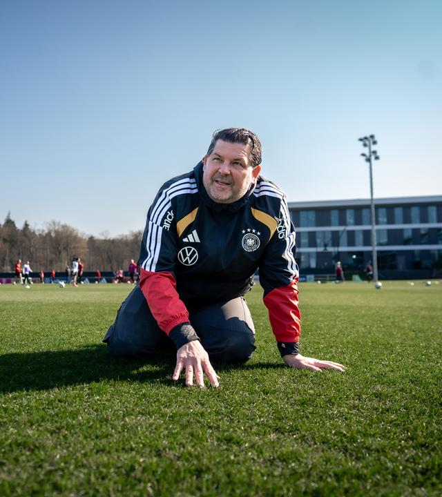Greenkeeper Sebastian Breuing kniet in schwarzer Jacke mit DFB-Logo auf einem Fußballfeld und stützt seine Hände auf den Rasen.