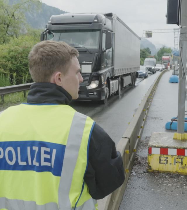 Zwei Polizisten mit gelben Warnwesten stehen an Grenzübergang, im Hintergrund Lkw und Autos im Stau