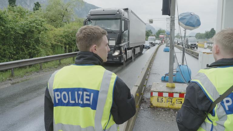 Zwei Polizisten mit gelben Warnwesten stehen an Grenzübergang, im Hintergrund Lkw und Autos im Stau