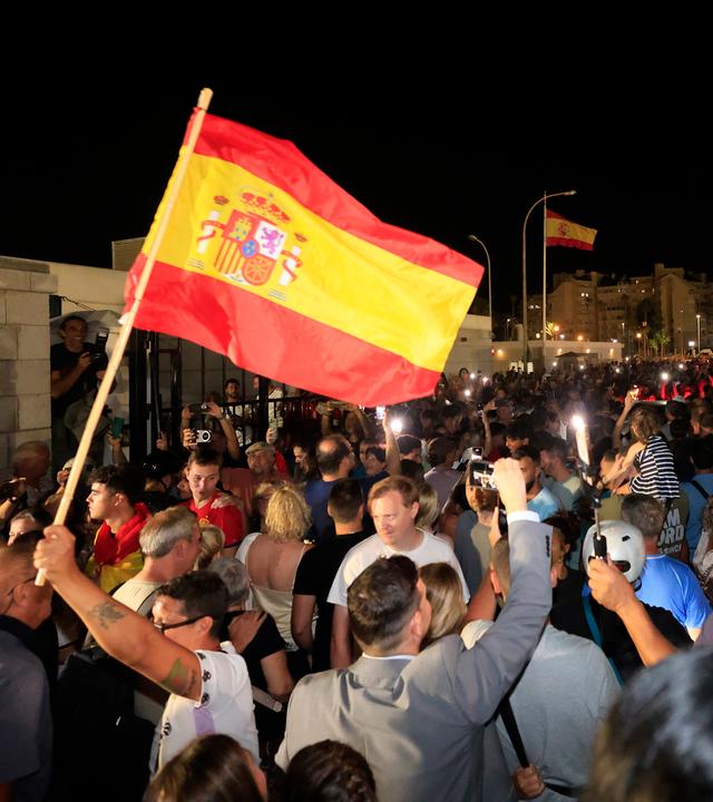 Jubelnde Menschen, die eine Spanien-Flagge schwenken stehen auf einer Straße, die vorher durch einen Grenzzaun getrennt war.
