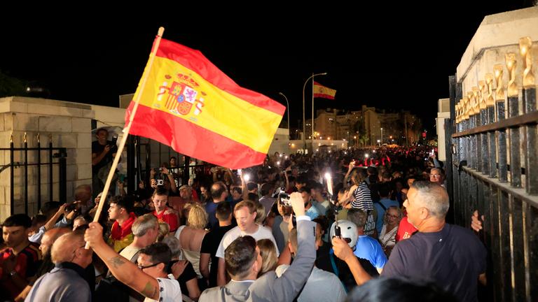 Jubelnde Menschen, die eine Spanien-Flagge schwenken stehen auf einer Straße, die vorher durch einen Grenzzaun getrennt war.