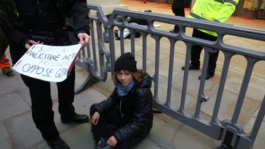 Greta Thunberg ist auf einer Demonstration. Sie sitzt auf dem Boden und neben ihr hält eine Person ein Plakat.