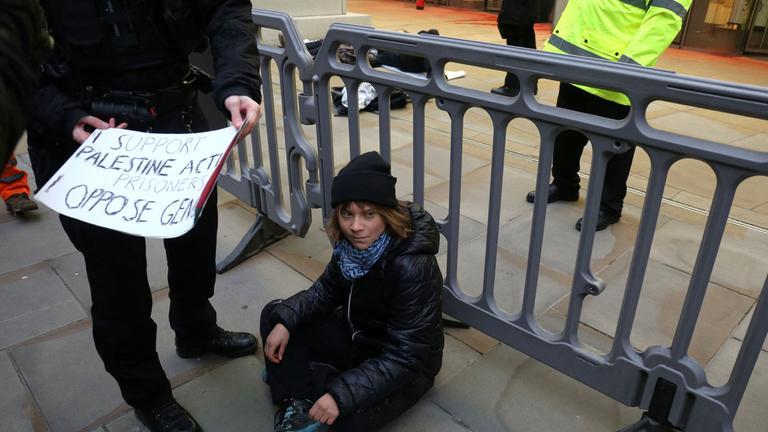 Greta Thunberg ist auf einer Demonstration. Sie sitzt auf dem Boden und neben ihr hält eine Person ein Plakat.