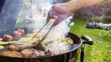 Auf dem Rost eines Grills liegen Steaks vom Rind, Lammkeule, Schweinenacken sowie Tomaten, Mais und Zucchini