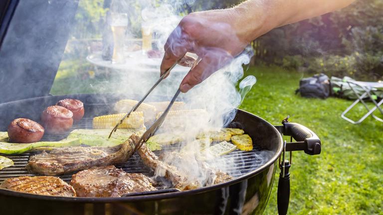 Auf dem Rost eines Grills liegen Steaks vom Rind, Lammkeule, Schweinenacken sowie Tomaten, Mais und Zucchini