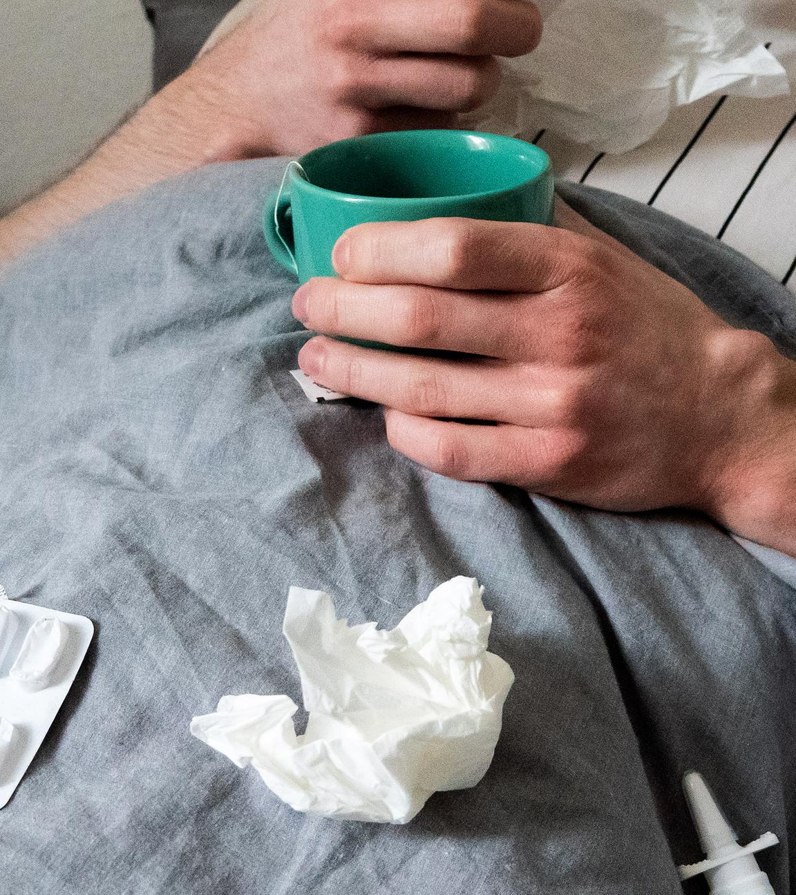 Symbolbild: Ein Mann liegt mit Taschentüchern, Teetasse, Nasenspray und Tabletten im Bett (gestellte Szene).